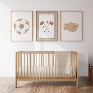 May include: Three framed prints with a soccer theme hang above a wooden crib. The prints feature a soccer ball, crossed flags with the letters M, A, and L, and a pair of soccer cleats. The crib has a beige blanket with white polka dots.