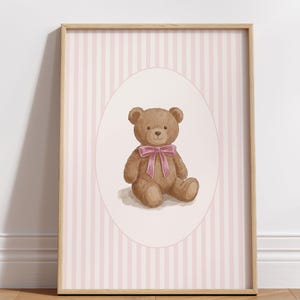 Puede incluir: Una impresión enmarcada con una ilustración en acuarela de un oso de peluche con un lazo rosa. El oso está sobre un fondo de rayas rosas y blancas dentro de un marco ovalado. La obra de arte se muestra en un marco de madera clara.