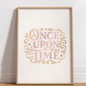 May include: A framed print with the words "ONCE UPON A TIME" in pink, surrounded by gold decorative flourishes. The print is in a light wood frame and leans against a white wall.