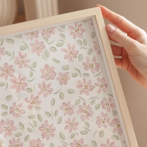 May include: A framed floral print featuring a pattern of pink flowers and green leaves on a white background. The frame is made of light-colored wood. The image is held by a hand.