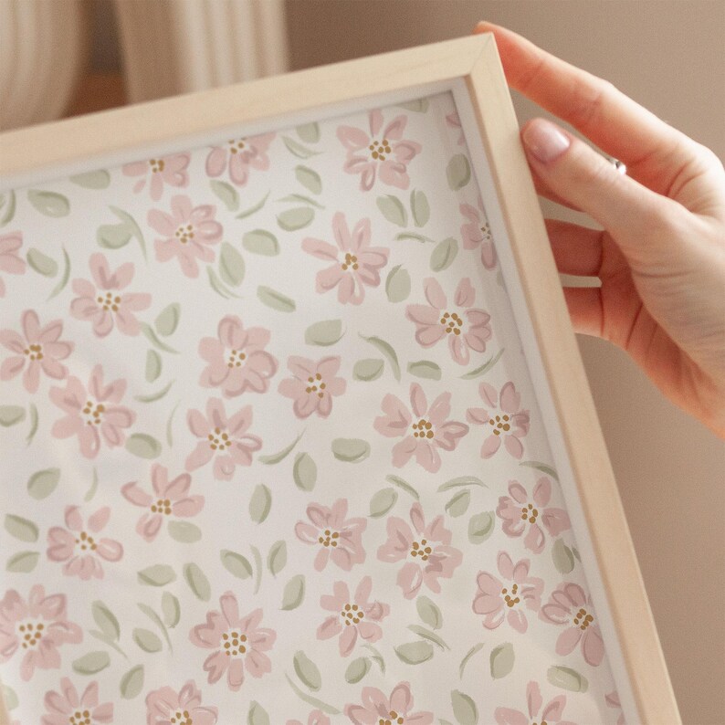 May include: A framed floral print featuring a pattern of pink flowers and green leaves on a white background. The frame is made of light-colored wood. The image is held by a hand.