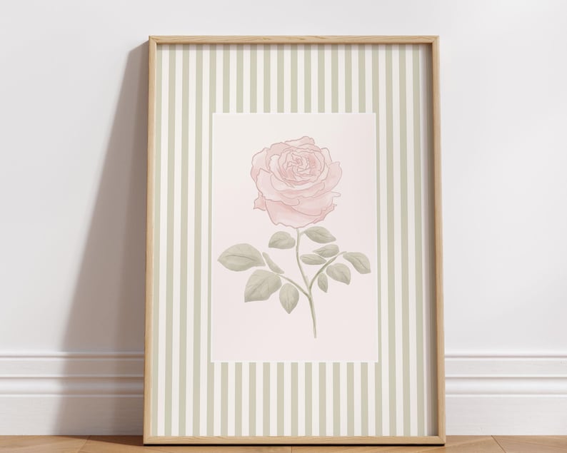 K&ouml;nnte beinhalten: Ein gerahmter Kunstdruck mit einer zarten rosa Rose mit gr&uuml;nen Bl&auml;ttern und Stiel. Das Kunstwerk ist vor einem hellrosa Hintergrund platziert und von einem hellgr&uuml;nen und wei&szlig;en gestreiften Rahmen umgeben, der in einem Holzrahmen eingefasst ist.
