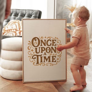 May include: A framed canvas print with the words "ONCE UPON A TIME" in a decorative gold font. The print is held by a child in a light-colored room. The print has a neutral background and a wooden frame.