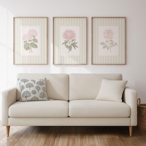 K&ouml;nnte beinhalten: Ein cremefarbenes Sofa mit einem dekorativen Kissen und drei gerahmten Blumendrucken. Die Drucke zeigen rosa Blumen auf gestreiftem Hintergrund. Ein Weidenkorb steht rechts auf dem Boden.