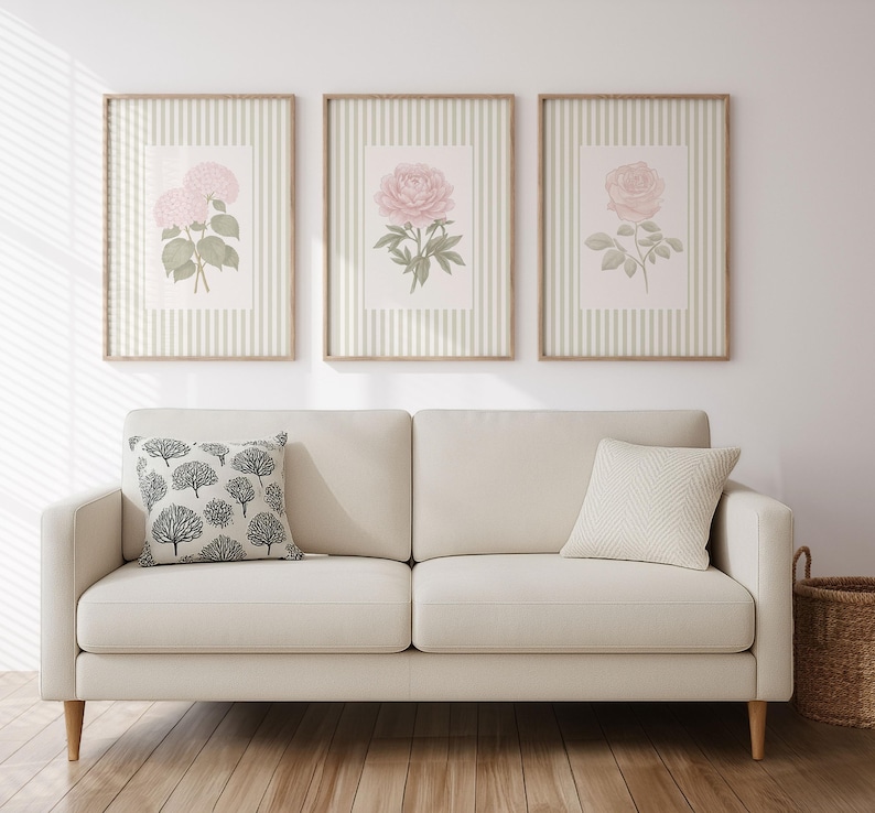 K&ouml;nnte beinhalten: Ein cremefarbenes Sofa mit einem dekorativen Kissen und drei gerahmten Blumendrucken. Die Drucke zeigen rosa Blumen auf gestreiftem Hintergrund. Ein Weidenkorb steht rechts auf dem Boden.