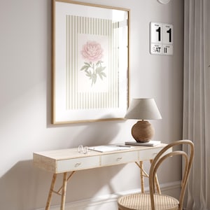 K&ouml;nnte beinhalten: Ein gerahmter Blumendruck mit einer rosa Pfingstrose auf gestreiftem Hintergrund h&auml;ngt &uuml;ber einem hellen Holzschreibtisch. Eine Schreibtischlampe mit beigem Schirm und Holzfu&szlig; steht auf dem Schreibtisch, zusammen mit einem Notizbuch und einer Brille. Ein Holzstuhl mit geflochtener Sitzfl&auml;che steht vor dem Schreibtisch.