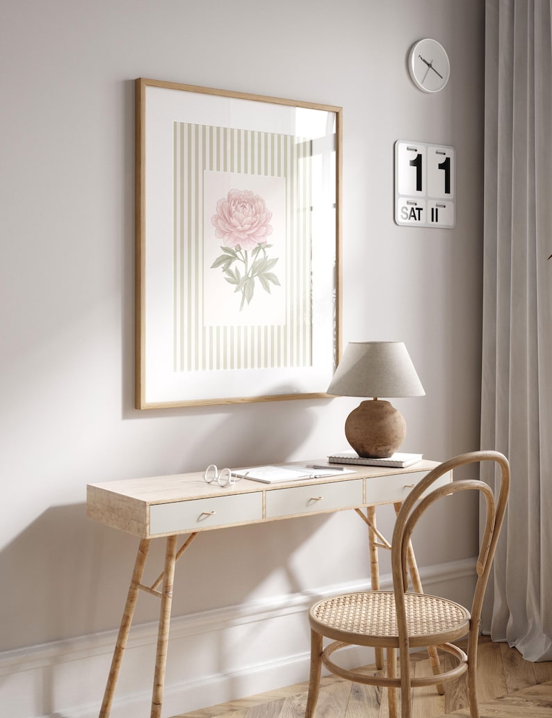 K&ouml;nnte beinhalten: Ein gerahmter Blumendruck mit einer rosa Pfingstrose auf gestreiftem Hintergrund h&auml;ngt &uuml;ber einem hellen Holzschreibtisch. Eine Schreibtischlampe mit beigem Schirm und Holzfu&szlig; steht auf dem Schreibtisch, zusammen mit einem Notizbuch und einer Brille. Ein Holzstuhl mit geflochtener Sitzfl&auml;che steht vor dem Schreibtisch.