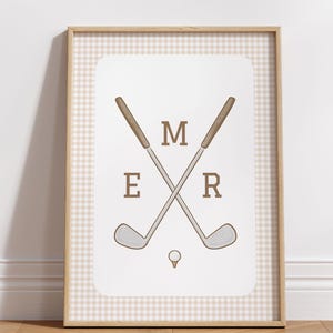 May include: Framed golf-themed art print. The artwork features two crossed golf clubs with the letters "MER" in a brown font. A golf ball is positioned below the clubs. The background is a white rectangle with a tan and white checkered pattern.