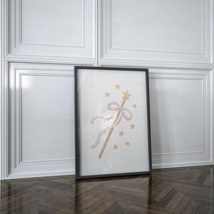 May include: A framed print featuring a whimsical illustration of a magic wand with a gold star, pink ribbon, and gold stars. The artwork is set against a white background, framed in black, and leans against a white wall.