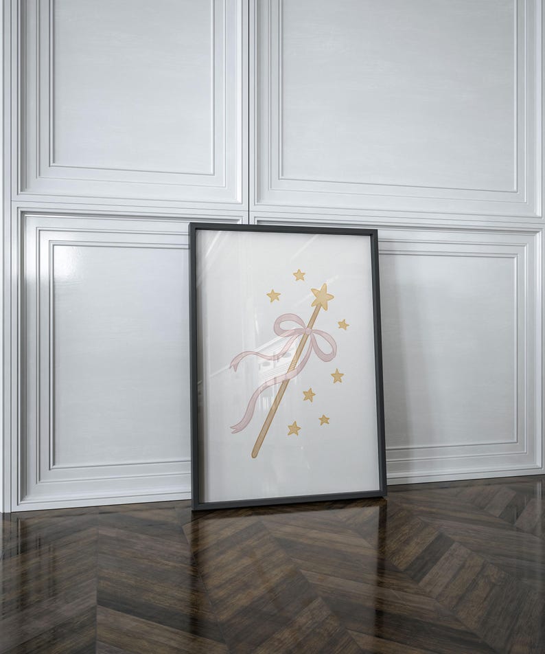 May include: A framed print featuring a whimsical illustration of a magic wand with a gold star, pink ribbon, and gold stars. The artwork is set against a white background, framed in black, and leans against a white wall.
