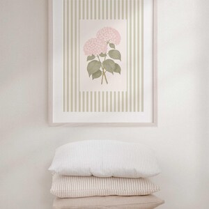 May include: A framed botanical print featuring pink hydrangea flowers with green leaves, set against a striped background. The frame is light-colored, and the print is displayed above a stack of four pillows in neutral tones.