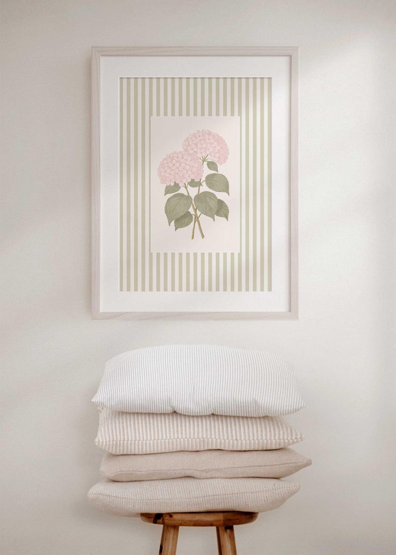 May include: A framed botanical print featuring pink hydrangea flowers with green leaves, set against a striped background. The frame is light-colored, and the print is displayed above a stack of four pillows in neutral tones.