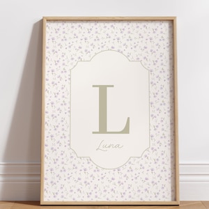 May include: A framed print with a floral pattern background and a central cream panel. The panel features a large, olive-green letter "L" and the name "Luna" in script. The frame is a light wood colour.
