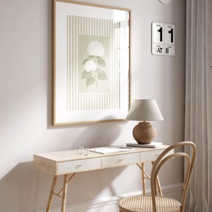 May include: A light wood desk with three drawers, a woven chair, and a framed botanical print. A beige lamp with a round base sits on the desk. A wall clock and a calendar are mounted on the wall.