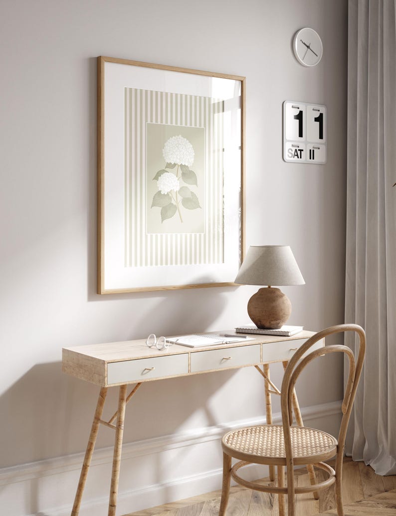 May include: A light wood desk with three drawers, a woven chair, and a framed botanical print. A beige lamp with a round base sits on the desk. A wall clock and a calendar are mounted on the wall.