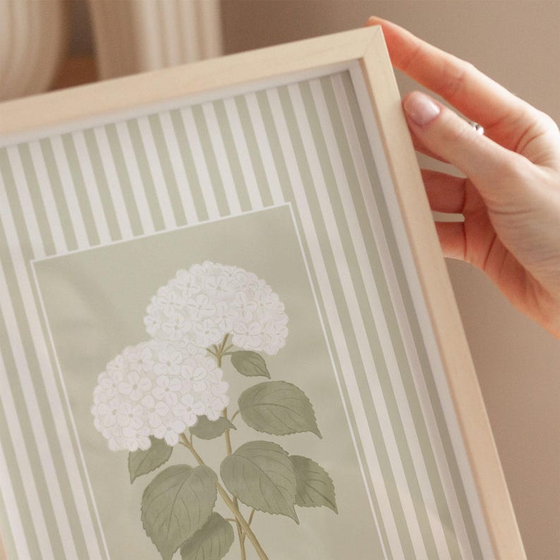 May include: A framed print featuring a botanical illustration of white hydrangea flowers with green leaves. The print is set against a light green background with vertical white stripes, encased in a light wood frame.