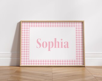 May include: A framed print featuring the name "Sophia" in pink, set against a white background. The frame has a pink and white gingham pattern. The artwork is displayed on a wooden floor against a white wall.