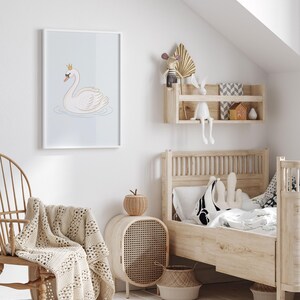 May include: A framed illustration of a white swan wearing a gold crown, floating on water. The artwork is displayed in a child's bedroom with a wooden bed, chair, and side table. A woven blanket and basket add to the room's decor.