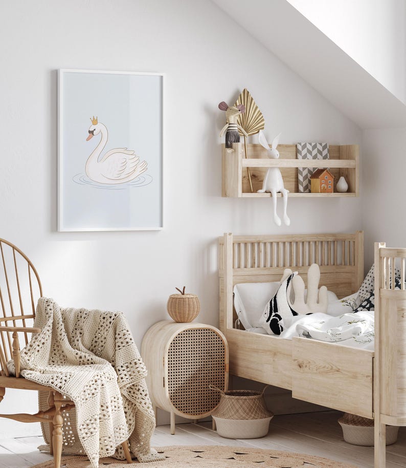 May include: A framed illustration of a white swan wearing a gold crown, floating on water. The artwork is displayed in a child's bedroom with a wooden bed, chair, and side table. A woven blanket and basket add to the room's decor.