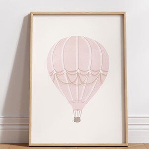May include: A framed print featuring a pink hot air balloon illustration. The balloon has a striped design with tan accents and a woven basket. The artwork is set against a light background, framed in natural wood.