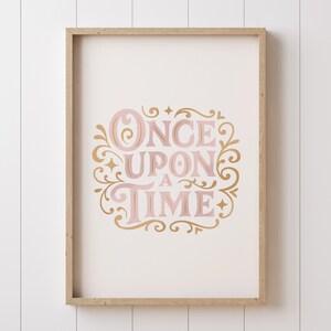 May include: A framed print with the words "ONCE UPON A TIME" in pink and gold script. The text is surrounded by decorative flourishes and set against a cream background. The frame is a light wood color.