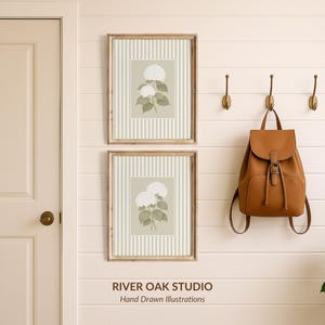 May include: Two framed botanical prints with white flowers and green leaves hang on a cream-colored wall. A brown leather backpack hangs from a hook. The text "RIVER OAK STUDIO Hand Drawn Illustrations" is at the bottom.