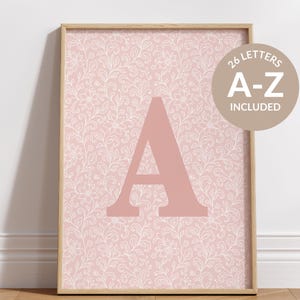 May include: A framed print with a large, pale pink letter "A" set against a pink backdrop with a white floral design. A circular sticker in the top right corner reads "26 LETTERS A-Z INCLUDED". The frame is a light wood colour.