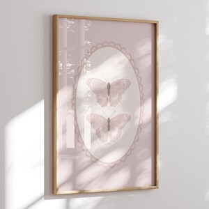 May include: A framed print featuring two butterflies in a soft pink oval frame. The butterflies are a muted pink color with detailed wing patterns. The background is a solid pink, and the frame is a light wood color.