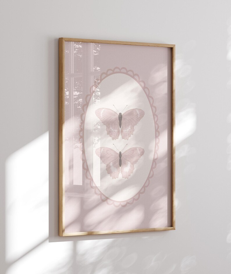 May include: A framed print featuring two butterflies in a soft pink oval frame. The butterflies are a muted pink color with detailed wing patterns. The background is a solid pink, and the frame is a light wood color.