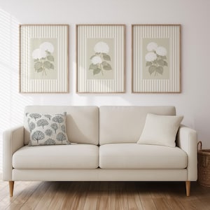 May include: Three framed botanical prints with white hydrangea flowers on a striped background hang above a cream-colored sofa. A decorative pillow with a tree pattern and a solid cream pillow rest on the sofa. A woven basket sits on the floor.