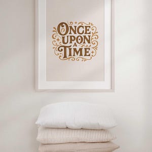 May include: A framed print with the words "ONCE UPON A TIME" in elegant, gold-colored script. Below the framed print, a stack of four pillows in varying neutral tones and textures rests on a wooden stool.
