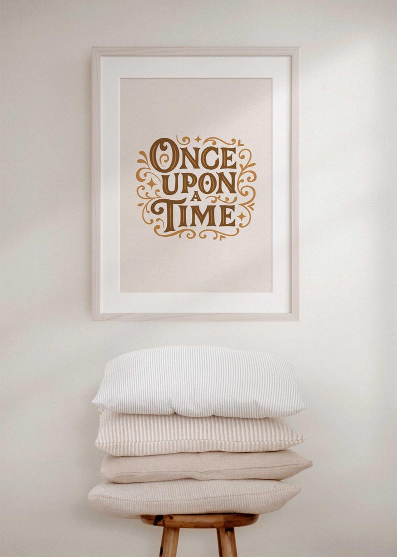May include: A framed print with the words "ONCE UPON A TIME" in elegant, gold-colored script. Below the framed print, a stack of four pillows in varying neutral tones and textures rests on a wooden stool.