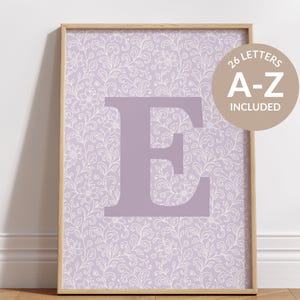 May include: A framed print featuring a large letter "E" in a soft purple hue. The background showcases a delicate white floral pattern on a lavender backdrop. A circular graphic states "26 LETTERS A-Z INCLUDED".