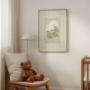 May include: A framed botanical print featuring white hydrangea flowers with green leaves. The artwork is set against a light green background with a striped border. The frame is a light wood color. The image is displayed in a room with a teddy bear and a chair.