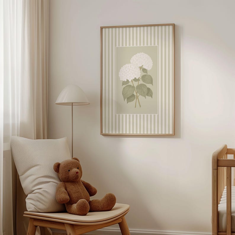 May include: A framed botanical print featuring white hydrangea flowers with green leaves. The artwork is set against a light green background with a striped border. The frame is a light wood color. The image is displayed in a room with a teddy bear and a chair.
