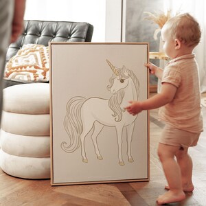 May include: A framed canvas art print featuring a white unicorn with a golden horn and hooves. The unicorn is depicted in a graceful pose against a neutral background. The artwork is displayed in a light-colored wooden frame, suitable for a nursery or children's room.