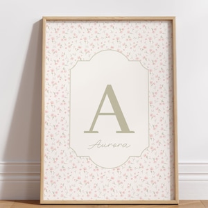 Op de afbeelding: Een ingelijste bloemenprint met een patroon van kleine roze bloemen op een lichtbeige achtergrond. Een centrale beige vorm toont de letter "A" en de naam "Aurora" in script. De lijst is licht hout.