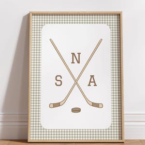 May include: A framed print with a gingham background featuring two crossed hockey sticks with the letters "N" and "A" on either side of the sticks and the letter "S" below the sticks. A small brown hockey puck is centered below the sticks.