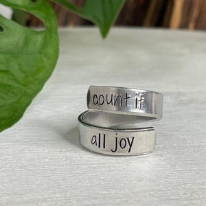 May include: A silver-colored wrap ring with the words "count it" and "all joy" stamped on the surface. The ring is displayed on a white surface with green foliage in the background.