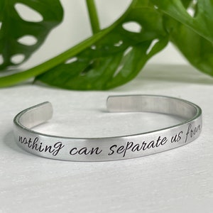 May include: A silver-toned metal cuff bracelet with an open back design. The exterior surface of the bracelet features the engraved black text "nothing can separate us from" written in a cursive script font along the length of the band.