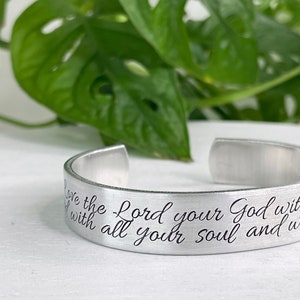 Puede incluir: Una pulsera de puño plateada con texto grabado. La pulsera abierta presenta la inscripción "Ama al Señor tu Dios con todo tu corazón y con toda..." en cursiva. El fondo incluye follaje verde.