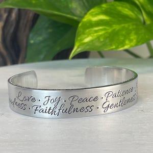May include: A silver-toned metal cuff bracelet featuring a brushed finish and black engraved text. The words Love, Joy, Peace, Patience, Faithfulness, and Gentleness are inscribed in a cursive script along the outer surface of the band.