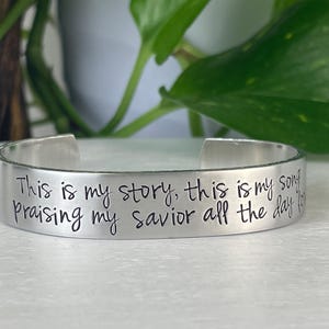 Puede incluir: Una pulsera de puño plateada con la inscripción "This is my story, this is my song, praising my savior all the day long." La pulsera abierta está sobre una superficie blanca, con follaje verde en el fondo.