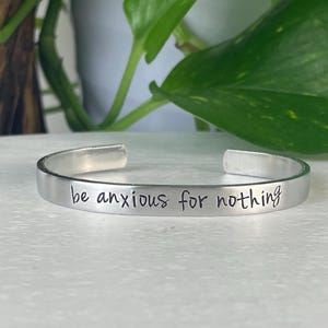 May include: A silver cuff bracelet with the phrase "be anxious for nothing" engraved in a handwritten style. The bracelet is displayed on a white surface with green foliage in the background.