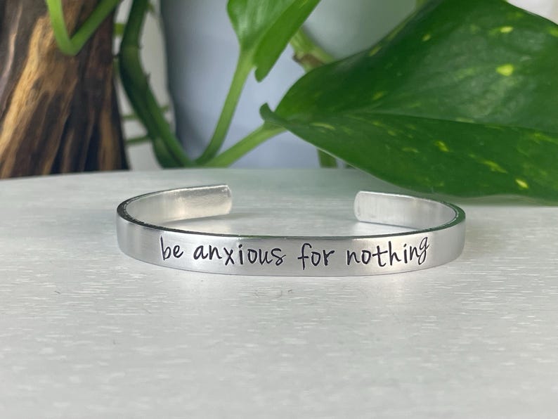 May include: A silver cuff bracelet with the phrase "be anxious for nothing" engraved in a handwritten style. The bracelet is displayed on a white surface with green foliage in the background.