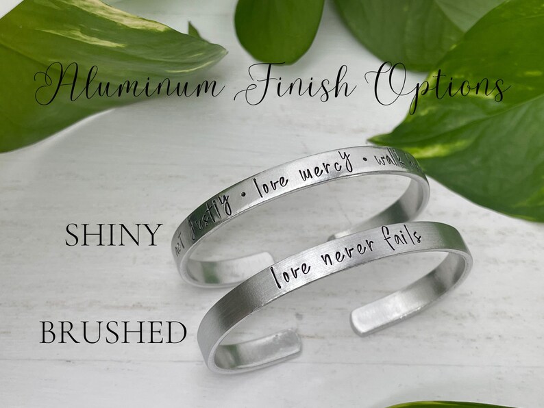 May include: Two silver aluminum cuff bracelets. One is shiny, the other brushed. The shiny bracelet has the words "not justify &bull; love mercy &bull; walk" engraved on it. The brushed bracelet has the words "love never fails" engraved on it. The text "Aluminum Finish Options" is at the top.