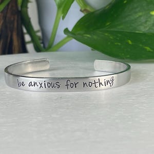May include: A silver cuff bracelet with the phrase "be anxious for nothing" engraved in a handwritten style. The bracelet is open-ended and sits on a white surface, with green foliage in the background.