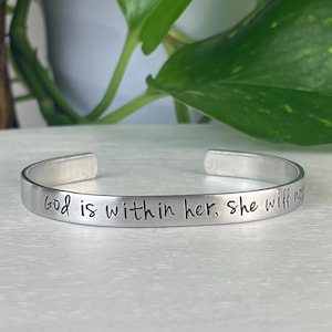 Puede incluir: Una pulsera de puño plateada con la inscripción "God is within her, she will not fail." La pulsera está hecha de metal liso y brillante y tiene un diseño abierto. El texto está en una fuente simple y elegante.