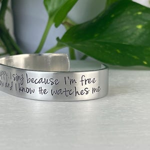 K&ouml;nnte beinhalten: Ein silberfarbenes Armreif-Armband mit eingraviertem Text. Der Text lautet "happy, I sing because I'm free, and I know He watches me." Das Armband wird auf einer wei&szlig;en Oberfl&auml;che mit gr&uuml;nem Laub im Hintergrund gezeigt.