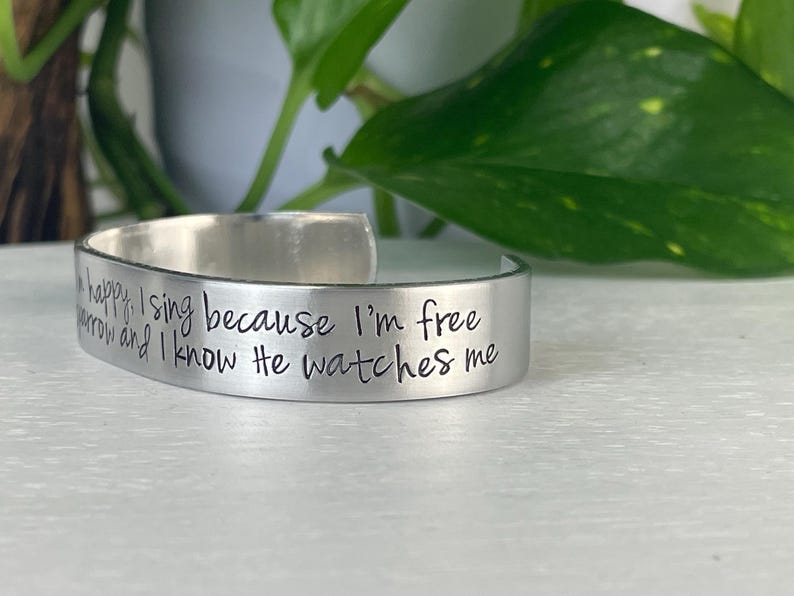 K&ouml;nnte beinhalten: Ein silberfarbenes Armreif-Armband mit eingraviertem Text. Der Text lautet "happy, I sing because I'm free, and I know He watches me." Das Armband wird auf einer wei&szlig;en Oberfl&auml;che mit gr&uuml;nem Laub im Hintergrund gezeigt.